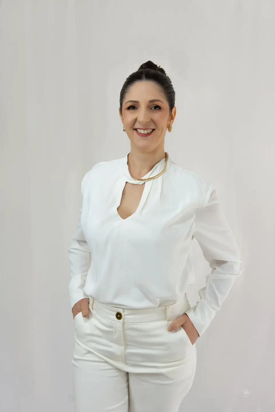 Retrato de Flávia Sangiovo sorrindo, de camisa e calça brancas, em estúdio de fundo claro