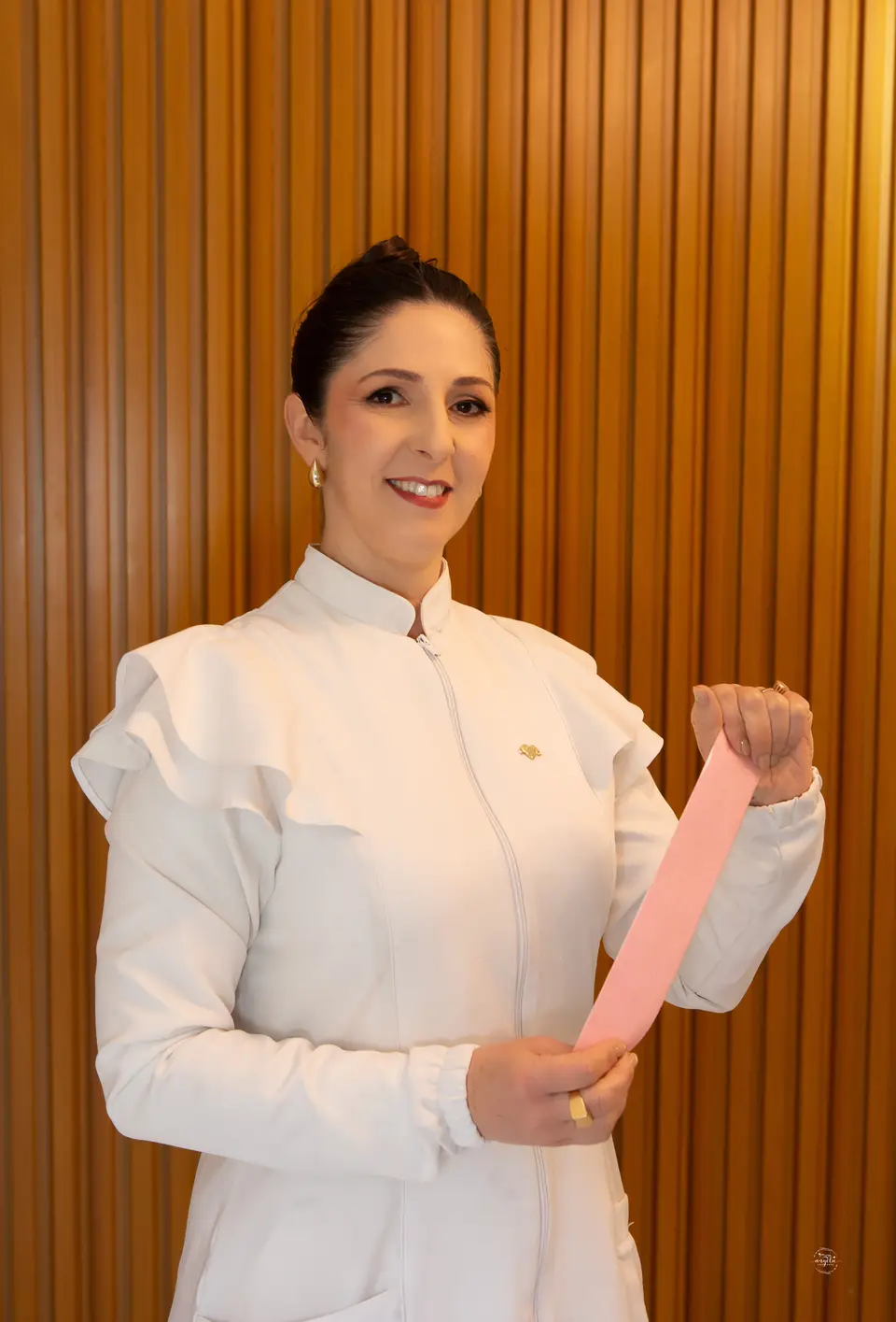 Flávia Sangiovo de jaleco branco segurando uma faixa de taping rosa, diante de um painel de madeira